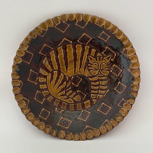 Puede incluir: Un plato redondo de cerámica con un diseño de gato marrón y beige. El gato está acurrucado en el centro del plato, rodeado por un fondo marrón oscuro con patrones de diamantes. El borde del plato tiene un borde festoneado texturizado.