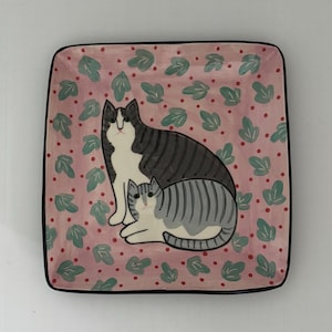 Puede incluir: Plato cuadrado de cerámica con fondo rosa con un estampado de hojas verdes y puntos rojos. El plato tiene un borde negro y representa dos gatos estilizados: uno sentado y otro tumbado, en tonos grises y blancos.