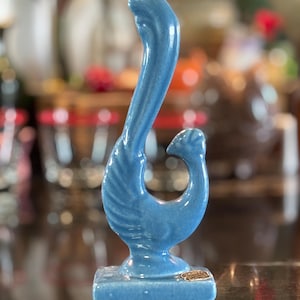 May include: A blue ceramic figurine shaped like a stylised bird with a long, curved neck and a small head. The bird is perched on a rectangular base and features a glossy finish. A small, rectangular sticker is visible on the front of the base.