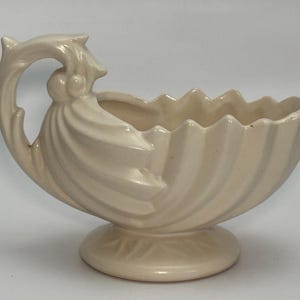 May include: Cream-colored ceramic vase with a shell-like design. The vase features a handle with a decorative flourish and a scalloped rim. It sits on a round base with a leaf pattern. The vase is approximately 8 inches long.