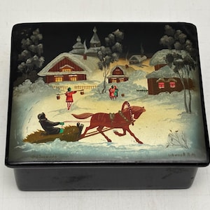 Puede incluir: Caja lacada en negro con una escena invernal pintada en la tapa. La escena representa un trineo tirado por caballos, casas y figuras en un paisaje nevado. La caja es rectangular y tiene un acabado brillante.