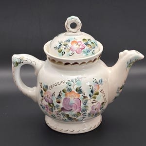 Könnte beinhalten: Cremefarbene Keramik-Teekanne mit Deckel, Henkel und Ausguss. Verziert mit floralen Designs in Rosa, Lila, Orange und Grün. Das Wort "сегодня" steht auf der Seite. Ein dekorativer Artikel für die Küche.