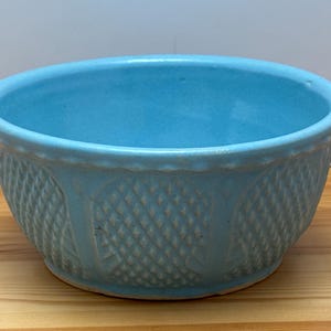 Puede incluir: Un cuenco de cerámica azul claro con un patrón texturizado similar a una cesta en el exterior. El cuenco tiene una forma redondeada y un interior liso. El cuenco está sobre una superficie de madera.