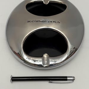 Peut inclure: Un cendrier chromé circulaire avec deux cavités noires et un repose-cigarette. Le mot "KOSMETIKA" est imprimé sur le cendrier. Un stylo noir avec une pointe et un clip argentés est à côté du cendrier.