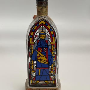 May include: A clear glass bottle with a stained-glass design depicting a figure in blue robes holding a yellow object. The design incorporates red, yellow, and blue colours. The bottle has a dark cap and a label at the base with the text "Vieille Cure".