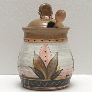 May include: A ceramic sugar bowl with a lid and a small spoon. The bowl is painted with a floral design in shades of pink, brown, and green. The lid and handle are brown, and the bowl has a black wavy pattern at the base.