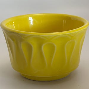 Könnte beinhalten: Eine leuchtend gelbe Keramikschale mit glänzender Oberfläche. Die Schale hat eine abgerundete Form mit einem gewellten Muster an der Außenseite. Die Innenseite ist glatt und die Schale steht auf einem kleinen, kreisförmigen Sockel.