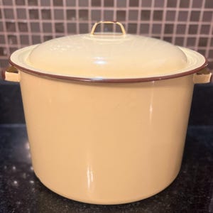 May include: A cream-colored enamel pot with a matching lid. The pot has a dark brown trim around the rim and a small handle on the lid. Two side handles are also present. The pot is sitting on a dark surface.