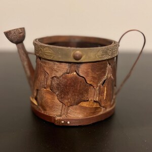 May include: A small, decorative watering can made of wood and metal. The body is constructed from overlapping pieces of brown wood, with a brass-colored metal band around the top. It has a small spout and a curved handle.