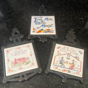 May include: Three vintage trivets with ceramic tiles and black metal frames. One tile reads "Free Milk with Meals," another "The Road to Friends House is Never Long," and the third "Give Us This Day Our Daily Bread."