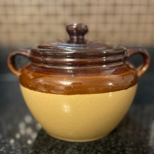 May include: A two-toned ceramic lidded jar with a round shape and two small handles. The jar is glazed in shades of brown and tan. The lid has a small knob for easy removal.