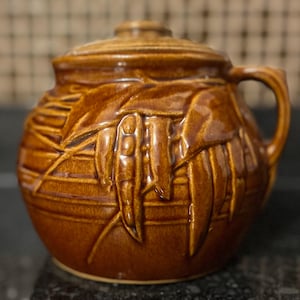 May include: A brown ceramic jar with a lid and handle. The jar features a textured design of leaves and pods. The jar is a warm brown color and appears to be glazed.