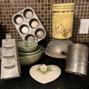 May include: A collection of vintage kitchen items, including a muffin tin, green bowls, a grater, a yellow canister with daisy design, a colander, a measuring sifter, and a heart-shaped dish with a daisy.