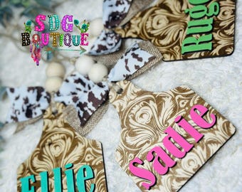 May include: Wooden cow tag ornaments with names in teal, green, and pink. The tags have a brown and cream floral pattern and are accented with cow print bows and wooden beads. The text "SDG Boutique" is also visible.