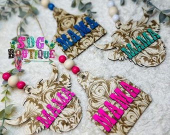 May include: Four wooden ornaments with a cow or bull head design. Each ornament has the word "MAMA" written on it in different colors. The ornaments are decorated with a brown and white swirl pattern.