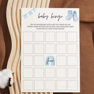 May include: A printable baby bingo card with a blue and white color scheme. The card has a free space with a rainbow and the text "free". The card has 25 squares for players to fill in with gifts they think will be opened at a baby shower.