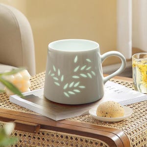 May include: A light gray ceramic mug with a curved handle and a leaf design. The mug is sitting on a magazine on a woven table. A small plate with a round pastry is in front of the mug.