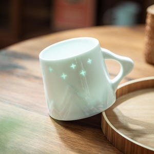 Może przedstawiać: Biały ceramiczny kubek z zakrzywionym uchwytem i świecącymi wzorami gwiazd. Kubek stoi na drewnianej powierzchni, z drewnianą tacą w tle. Kubek ma perłowe wykończenie.