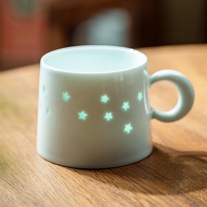 Könnte beinhalten: Eine weiße Keramik-Tasse mit rundem Henkel und sternförmigen Aussparungen. Die Aussparungen strahlen ein sanftes, grünes Leuchten aus. Die Tasse steht auf einer Holzoberfläche.