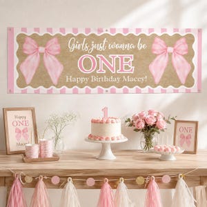 May include: A rectangular birthday banner with a tan background, pink striped borders, and two large pink bows. The text reads Girls just wanna be ONE Happy Birthday Macey! The banner is displayed above a table with a cake, flowers, and frames.