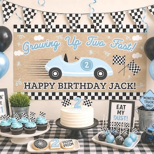 May include: A race car themed birthday party display featuring a banner with the text Growing Up Two Fast! Happy Birthday Jack!, light blue and black balloons, a white cake with a number two topper, cupcakes, and a blue toy monster truck.