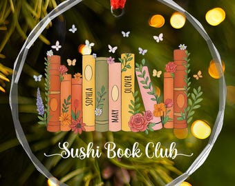 May include: A clear, round ornament with a red ribbon for hanging. The ornament features a stack of colorful books with floral and butterfly accents. The words "Sushi Book Club" are written in a script font.