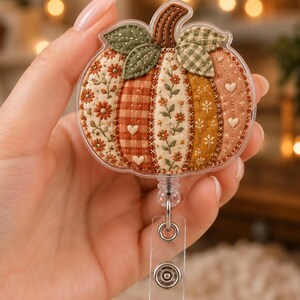 May include: A pumpkin-shaped badge reel featuring a quilted fabric design with alternating panels of floral, gingham, and solid autumn-toned fabrics. The pumpkin is topped with a brown stem and green leaves, encased in clear plastic with a metal clip.