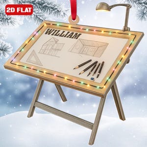 May include: A wooden artist's drafting table ornament with the name "WILLIAM" on the drawing surface. The table features a drawing of a house, pencils, and a small lamp. The frame is decorated with colorful lights, and a red ribbon is attached for hanging.