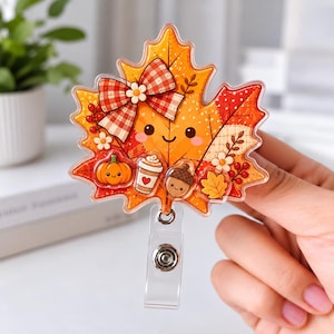 May include: An acrylic badge reel featuring a large, orange maple leaf with a smiling face. The leaf is decorated with a red and white plaid bow, a small orange pumpkin, a coffee cup, an acorn, and white flowers with red berries.