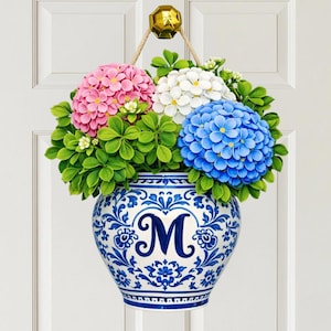May include: A decorative door hanging featuring a blue and white ceramic vase filled with pink, white, and blue hydrangea flowers and green leaves. The vase has a blue floral design and the letter "M". The hanging is suspended by a rope.