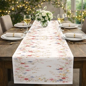May include: A white floral table runner with a delicate pattern of red, pink, blue, and yellow flowers and butterflies. The runner is placed on a dark wooden table set for a meal, with plates, silverware, and wine glasses.