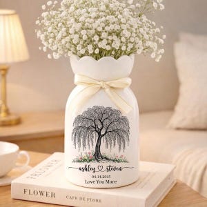 May include: White ceramic vase with a weeping willow tree design, a cream-colored ribbon, and a floral arrangement. The vase has the names "ashley ♡ steven" and the date 04.14.2015, with the words "Love You More".