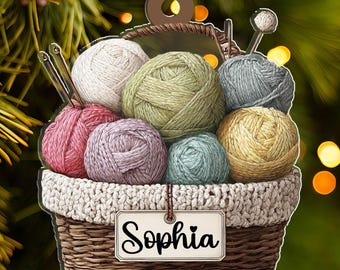 May include: A 2D flat ornament featuring a basket filled with colorful yarn balls and knitting needles. The basket has a woven design and a tag that reads "Sophia". The ornament is designed for hanging and is set against a blurred green background.