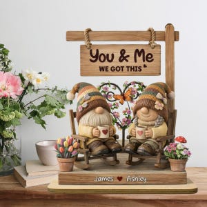 May include: A decorative wooden sign with the text "You & Me WE GOT THIS" above two gnome figurines. The gnomes are seated in rocking chairs, holding mugs with hearts. The base is personalized with names.