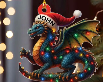 May include: A 2D flat Christmas ornament featuring a blue dragon wearing a Santa hat with the name "JOHN". The dragon is adorned with colorful Christmas lights. The ornament is held by a gold string.