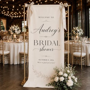 Puede incluir: Una pancarta vertical de tela color crema suspendida de un soporte de metal dorado con cintas beige. El texto dice: WELCOME TO Audrey's BRIDAL shower OCTOBER 8, 2026 We're so glad you're here! Una ilustración floral aparece en la parte inferior.