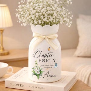 May include: A white ceramic vase with a scalloped rim, tied with a cream-colored ribbon, and filled with white flowers. The vase has the text "Chapter Forty" and "Anna" with butterfly and floral accents. The vase rests on a book.
