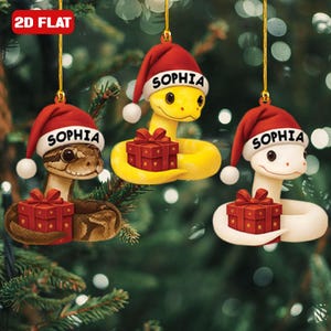 May include: Three Christmas ornaments shaped like snakes, each wearing a red Santa hat with the name "SOPHIA" on it. The snakes are in shades of brown, yellow, and white, and each holds a red gift box. The ornaments are hanging from gold strings.