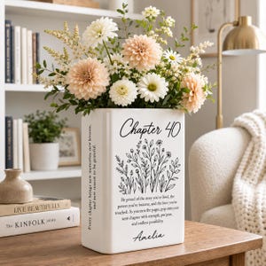 May include: A white rectangular ceramic vase featuring black text that reads Chapter 40, a floral line art illustration, and a personalized message for Amelia. The vase is filled with a bouquet of peach dahlias, white daisies, and green foliage.