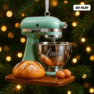 Puede incluir: Adorno navideño de batidora de pie verde menta con un cuenco plateado grabado con "Laura". También hay un pan, dos huevos y una tabla de cortar de madera. El adorno tiene una cinta blanca para colgar. El texto "2D FLAT" es visible.