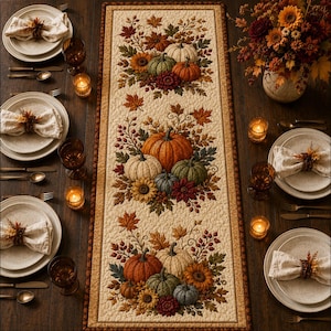 May include: A rectangular quilted table runner featuring a cream background with three repeating clusters of pumpkins, sunflowers, and autumn leaves in shades of orange, green, and brown, bordered by a dark brown trim on a wooden dining table.