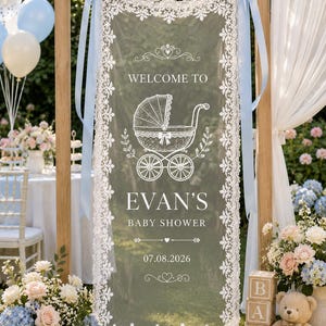 May include: A rectangular white lace banner featuring a central graphic of a vintage baby carriage. The text reads "WELCOME TO EVAN'S BABY SHOWER" with the date "07.08.2026" below. The banner is suspended by light blue ribbons from a wooden frame.