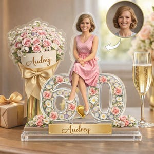 Puede incluir: Un adorno de pastel de acrílico personalizado con la figura de una mujer con vestido rosa sentada sobre un número 60 grande lleno de rosas rosas y margaritas blancas. Un ramo muestra el nombre Audrey y una placa dorada dice Audrey.