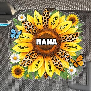 May include: A sunflower-shaped car accessory with the word "NANA" in white block letters. The sunflower has yellow petals with leopard print accents, and the names "Emma", "Emily", "Sarah", "Maria", "Jolee", "William", and "Elizabeth" are written on the petals. Butterflies and daisies are also included.