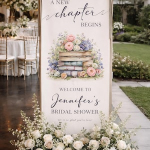 May include: A large sign with the text "A New Chapter Begins" and "Welcome to Jennifer's Bridal Shower". The sign features a watercolor illustration of books and flowers. A floral arrangement of white roses and greenery is at the base.
