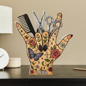 May include: A wooden hand-shaped organizer with a floral and butterfly design, holding a black comb, scissors, and shears. The base of the organizer has the name "Bella" written on it. The color scheme includes tan, black, red, and blue.