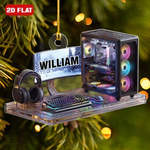 May include: A clear acrylic ornament featuring a gaming setup. It includes a computer, monitor with "WILLIAM" text, keyboard, mouse, and headphones. The computer has colorful internal lights. The ornament is labeled "2D FLAT".