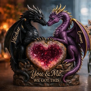 May include: A decorative sculpture featuring two dragons facing each other, flanking a heart-shaped geode. One dragon is black, the other purple, with gold accents. The geode is filled with sparkling red crystals. The base reads "You & Me WE GOT THIS."