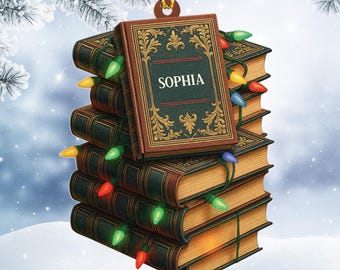 May include: A Christmas ornament shaped like a stack of books, with the name "SOPHIA" on the top book. The books are brown with gold accents, and colorful Christmas lights are strung around them. The ornament is hanging from a gold string.