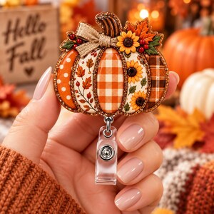 May include: A pumpkin-shaped badge reel featuring a multi-patterned design with orange polka dots, autumn leaves, orange gingham, sunflowers, and brown plaid. The top is accented with a tan burlap bow and a glittery border finish.